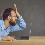 Frustrated businessman slapping his face at office desk after making a critical UI/UX mistake on his laptop.