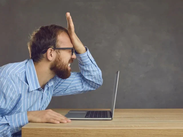 Frustrated businessman slapping his face at office desk after making a critical UI/UX mistake on his laptop.