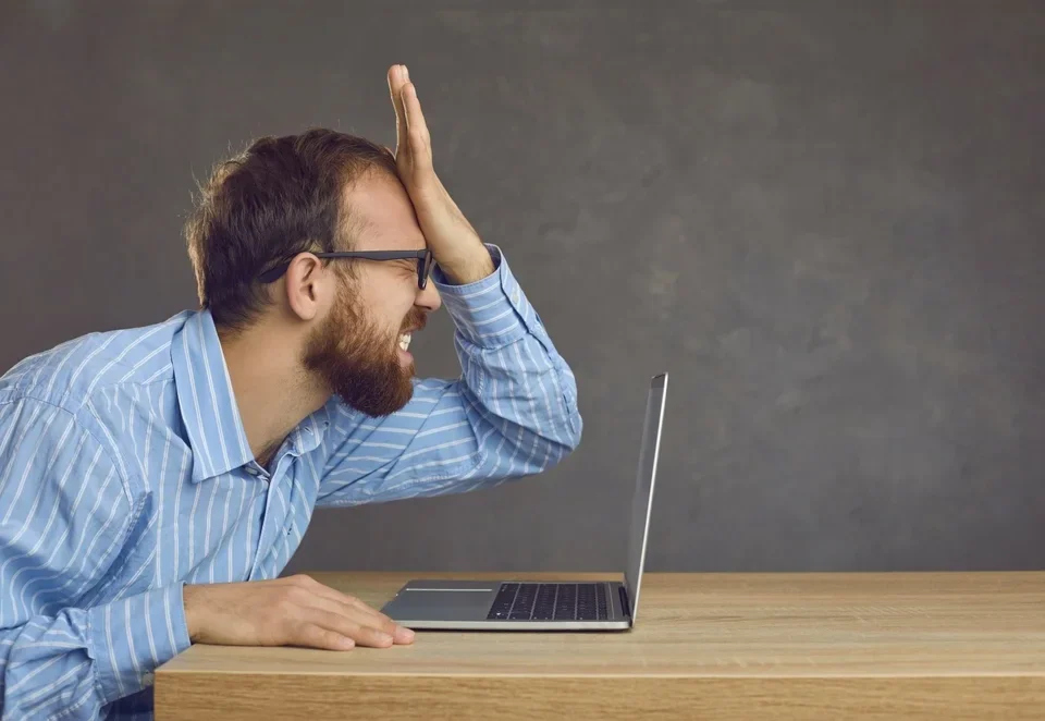 Frustrated businessman slapping his face at office desk after making a critical UI/UX mistake on his laptop.