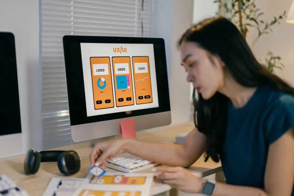 Asian female developer designing a mobile app in a cluttered workspace with code and wireframes on screen.