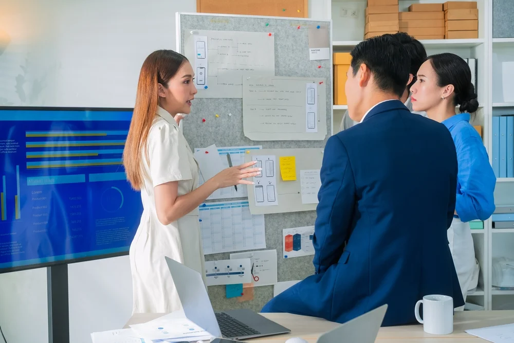 Asian UX strategist and programmer discussing user journey and feedback strategy in a modern digital workspace.