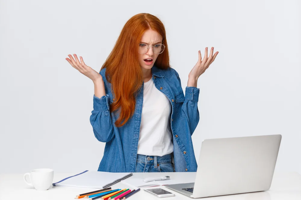 Frustrated redhead woman reacting to inconsistent user interface elements on her laptop.